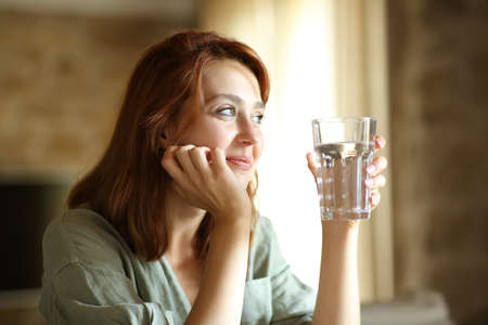 Satisfied woman holding water glass sitting at home contemplating through a windowの写真素材