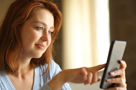 Satisfied redhead woman using smart phone sitting at homeの写真素材