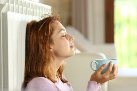 Profile of a woman relaxing and heating on radiator holding coffee cup at homeの写真素材