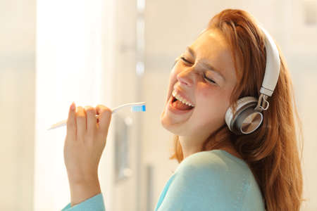 Happy lady listening to music with wireless headphones and singing using a toothbrush as a mic in the bathroomの写真素材