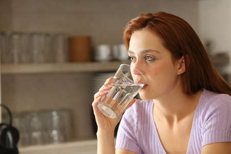 Serious woman drinking water from glass in the kitchen at homeの写真素材