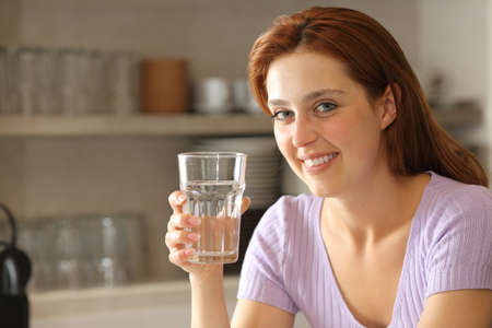 Happy woman holding water glass looking at camera in the kitchen at homeの写真素材