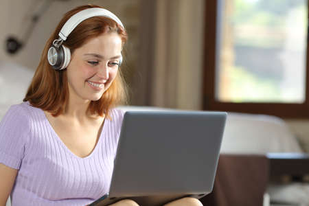 Happy woman using laptop and headphones to watch videos in a bedroomの写真素材