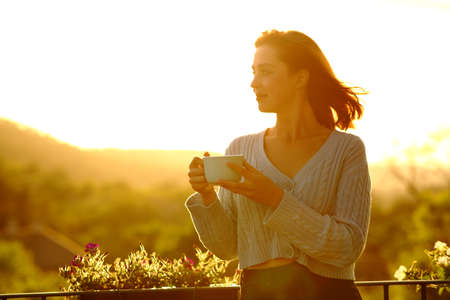 Confident woman holding coffee cup contemplating at sunset in a balconyの写真素材
