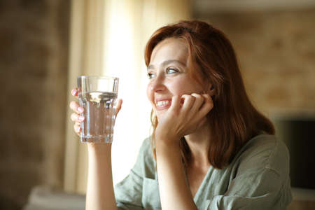 Happy woman holding water glass sitting in the living room at home looking awayの写真素材