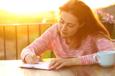 Concentrated woman writing on paper agenda at sunset in a balconyの写真素材
