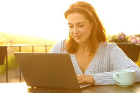 Relaxed woman is using laptop sitting in a balcony at sunsetの写真素材