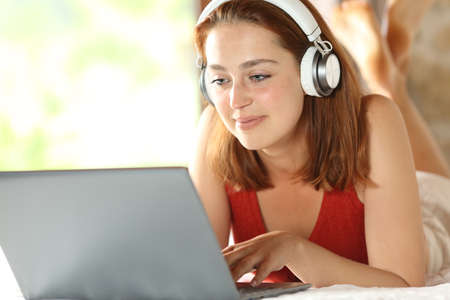 Satisfied woman with wireless headphones checking laptop lying on the bed in the bedroomの写真素材
