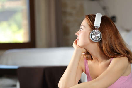 Serious woman relaxing listening to music with wireless headphones looking through a window in the bedroomの写真素材