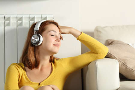 Relaxed woman sitting leaning on radiator listening to music with headphones at homeの写真素材