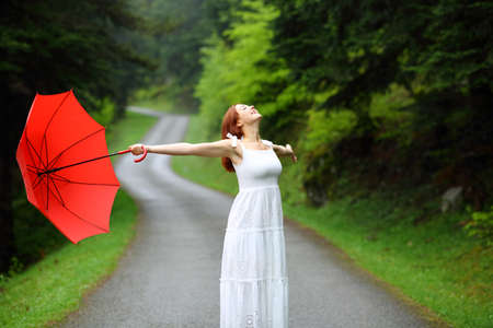Happy woman in white dress holding red umbrella enjoying under rain in a road of a green forestの写真素材