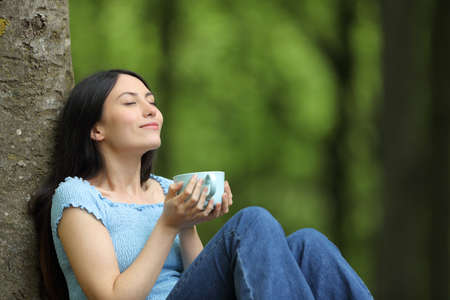 Happy asian woman drinking coffee relaxing breathing fresh air sitting in a forestの写真素材