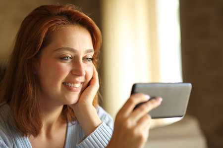 Happy woman watching media online content on smart phone at homeの写真素材