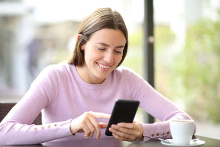 Happy woman using mobile phone checking online content sitting in a coffee shopの写真素材