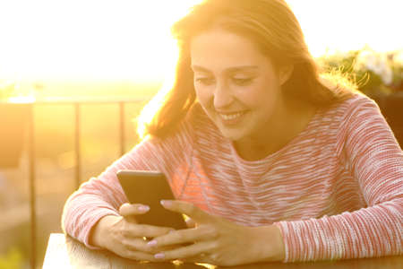Happy woman using smart phone sitting in a house balcony at sunset with a warm lightの写真素材