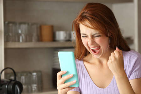 Excited woman checking smart phone content in the kitchen at homeの写真素材