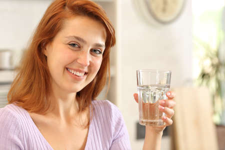 Happy female posing looking at camera holding a glass of water in the kitchenの写真素材