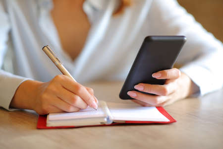 Close up of a businesswoman writing on agenda checking smart phone at officeの写真素材