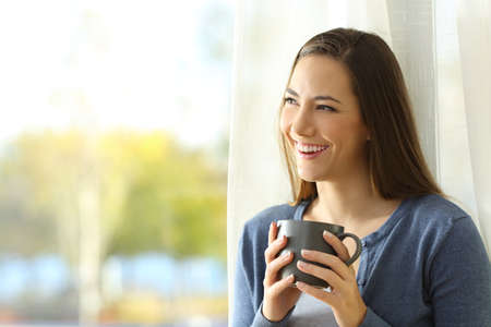 Happy pensive woman looks at side beside a window holding a coffee cup at homeの写真素材