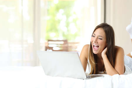 Happy woman watching online videos on a laptop lying on a bed at homeの写真素材