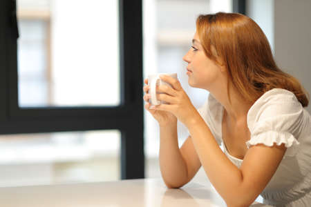 Pensive lady drinking coffee looking through a window at homeの写真素材
