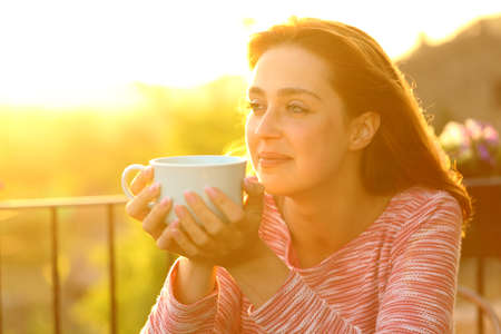 Satisfied woman contemplating views holding coffee mug at sunset in a balconyの写真素材
