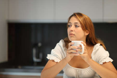Pensive woman looking away holing coffee cup in the kitchen at homeの写真素材
