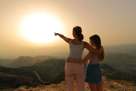 Two women pointing at sun at sunset in the mountainの写真素材