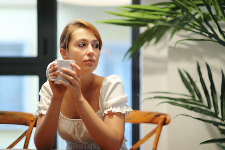 Serious woman drinking coffee thinking looking at side sitting on a chair at homeの写真素材