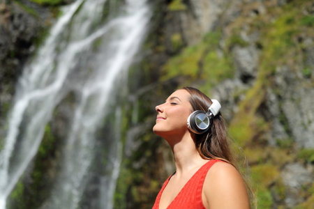 Happy woman in red listening to music wearing headphones breathing fresh air in a waterfallの写真素材