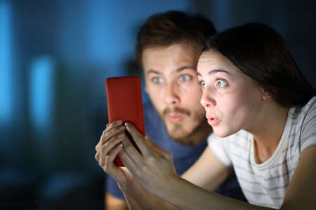 Surprised couple checking smart phone in the night at homeの写真素材