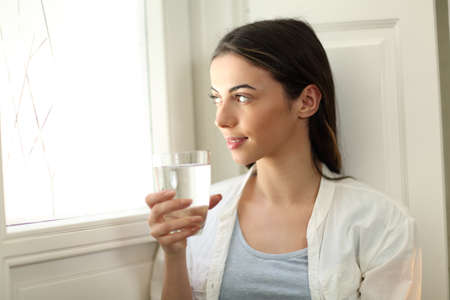 Happy woman holding water glass lokking through window sitting at homeの写真素材