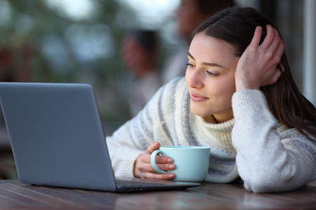 Happy woman holding coffee mug checking laptop content in winter in a terraceの写真素材