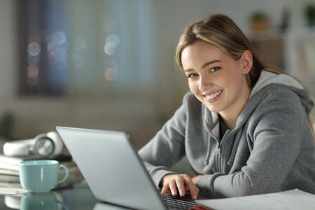 Happy student using laptop looking at you in the night at homeの写真素材