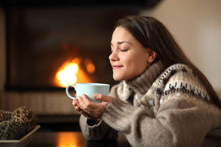 Happy woman in winter drinking coffee beside a fireplace heating at homeの写真素材