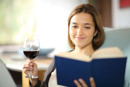 Happy woman reading a paper book drinking wine sitting on a couchの写真素材