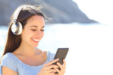 Happy woman listening to music wearing headphones and checking smart phone on summer vacatin on the beachの写真素材