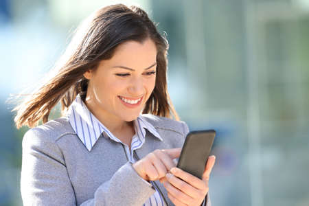 Happy businesswoman checking smart phone walking outdoors in the streetの写真素材