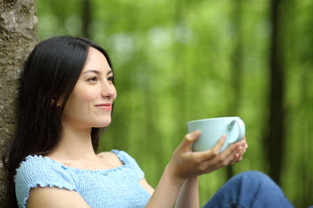 Happy asian woman looking away and drinking coffee sitting in a parkの写真素材