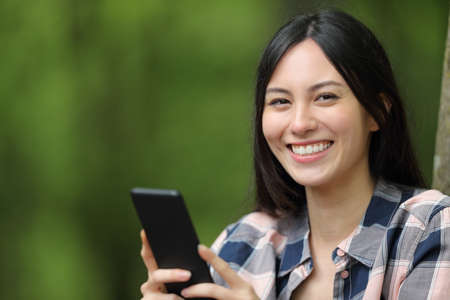 Asian woman smiling holding smart phone looking at camera in a parkの写真素材