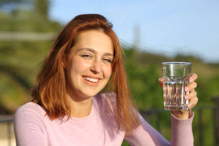 Happy woman holding glass of water smiling at camera in a balconyの写真素材