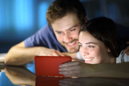 Happy couple watching videos on smart phone on a table in the night at homeの写真素材