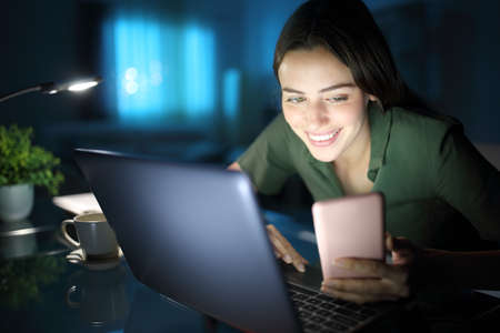 Happy woman in the night at home using laptop and checking smart phoneの写真素材