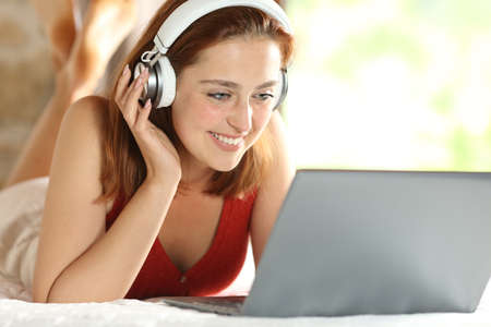 Happy woman wearing headphones watching media content on laptop on the bedの写真素材