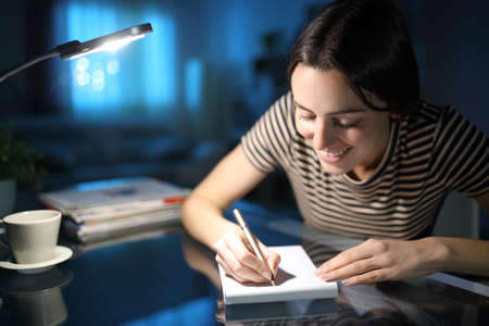 Happy woman taking notes on paper notebook in the night at homeの写真素材