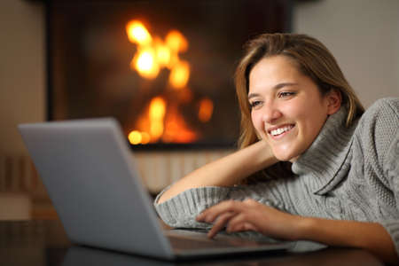 Happy woman using laptop watching media content at home near fireplaceの写真素材