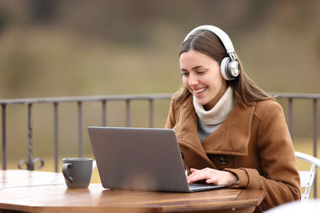 Happy woman in winter using laptop with headphones in a terraceの写真素材