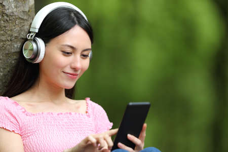 Asian woman listening to music checking smart phone sitting in a parkの写真素材
