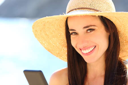 Happy woman holding smart phone looking at you smiling on the beachの写真素材