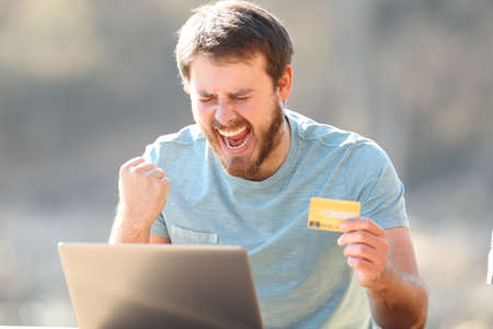 Excited man holding credit card buying online on laptop celebrating success outdoorsの写真素材